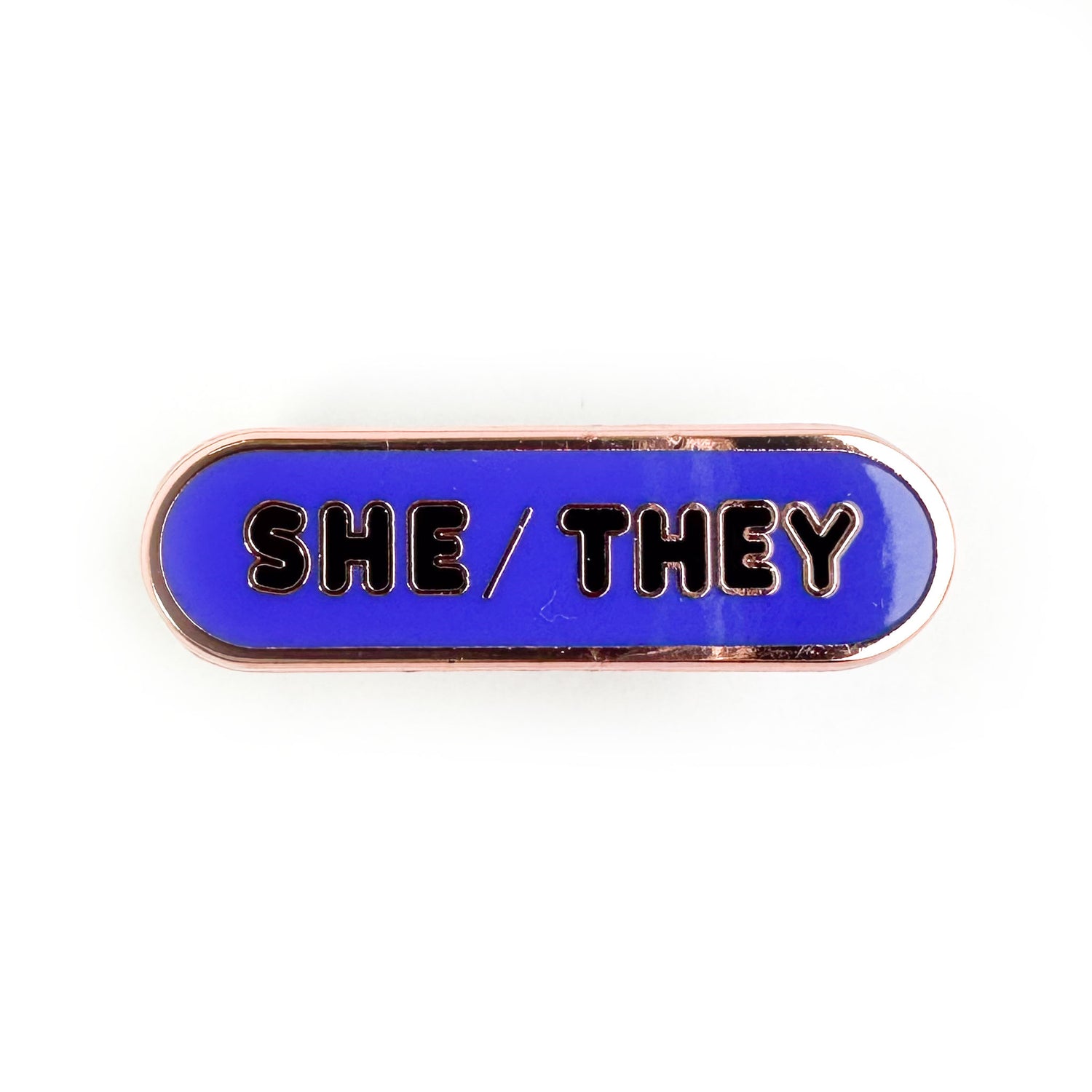 An indigo oval shaped pin with bubble letters on it that spell "She/They" on it. 