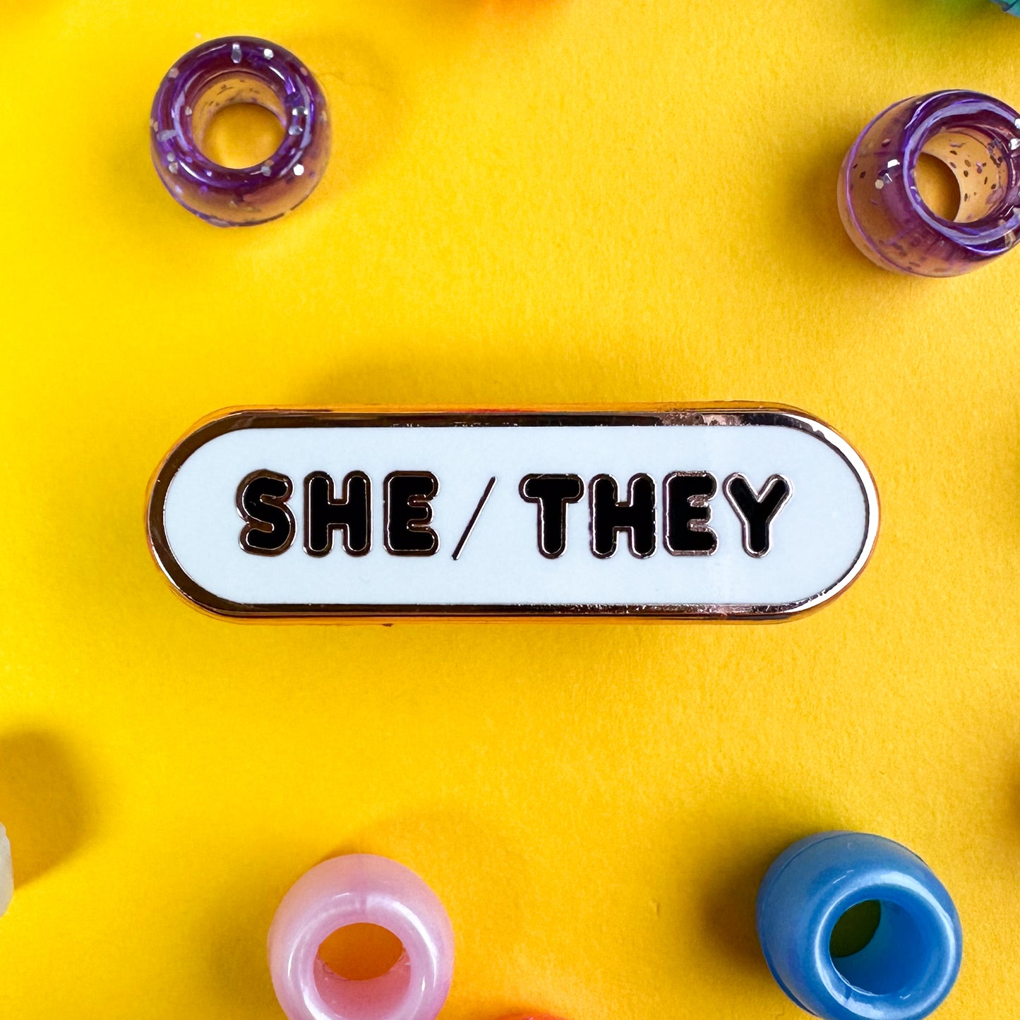 An oval shaped pin with the pronouns "She/They" on it in black letters. The pin is on a yellow background with purple, blue, and pink pony beads around it.