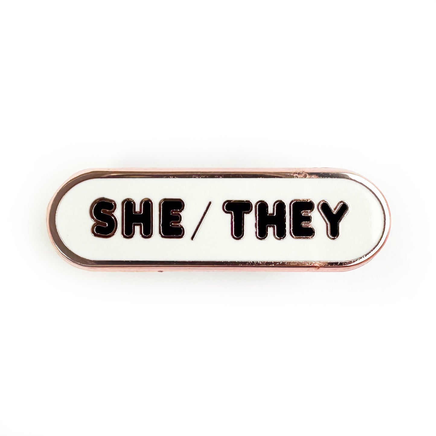 A pin with bubble letters that spell the pronouns "She/They" on it. 