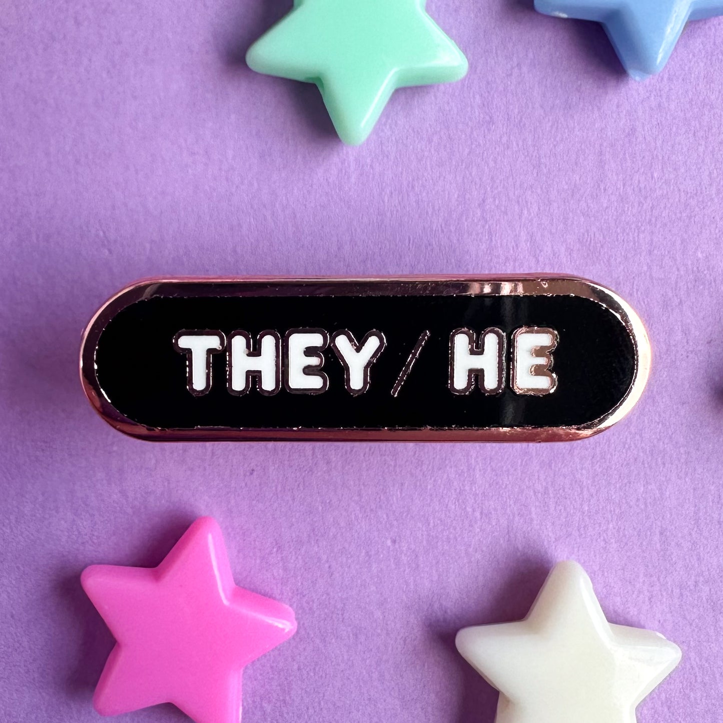 A capsule shaped enamel pin that is mostly black with white bubble letters. The pin is on a purple background with plastic star beads around it. 