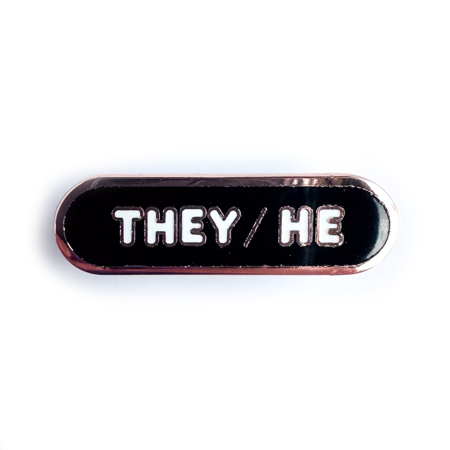 A black capsule shaped pin with copper metal and white bubble letters that have the pronouns They/He on it. 