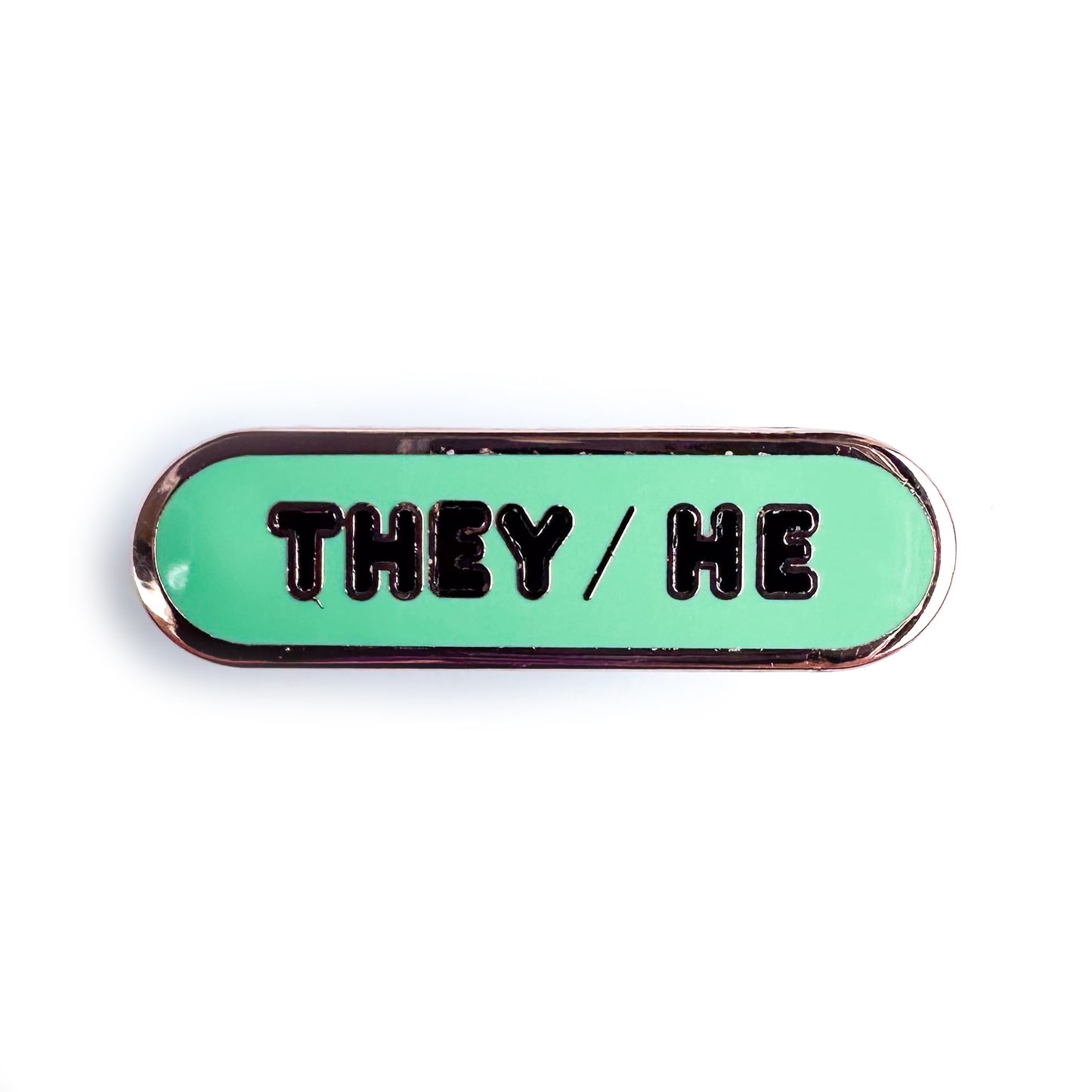 A capsule shaped pin that is green with black bubble letters with They/He on it. 