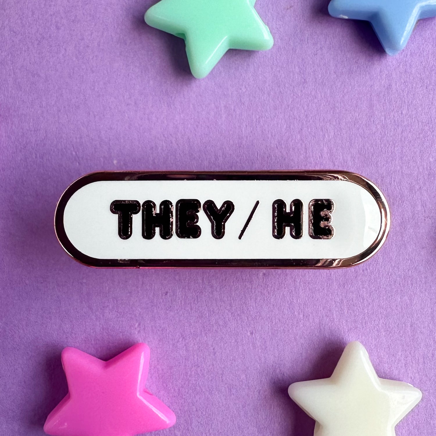 A white oval shaped enamel pin with black bubble letters with the pronouns "They/He". The pin is on a purple background with plastic star beads around it. 