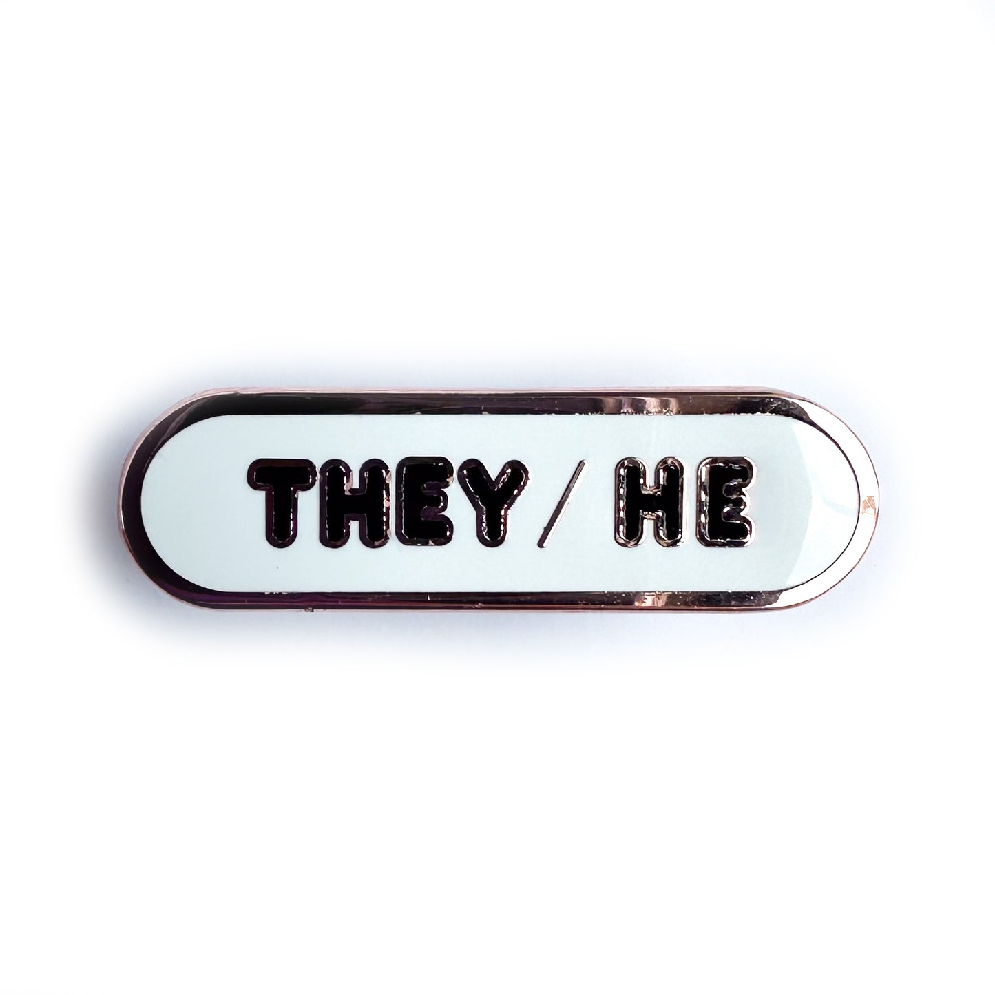 A white oval shaped pin with "They/He" on it in black bubble letters.