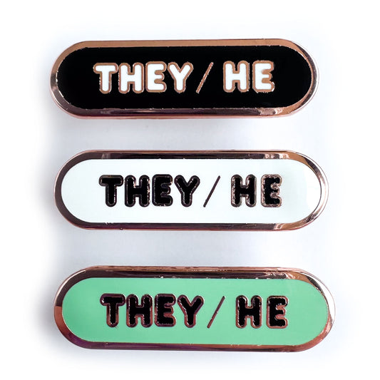 Three capsule shaped enamel pins with the words "They/He" on them, the top pin is black, the middle is white, and the bottom one is green.