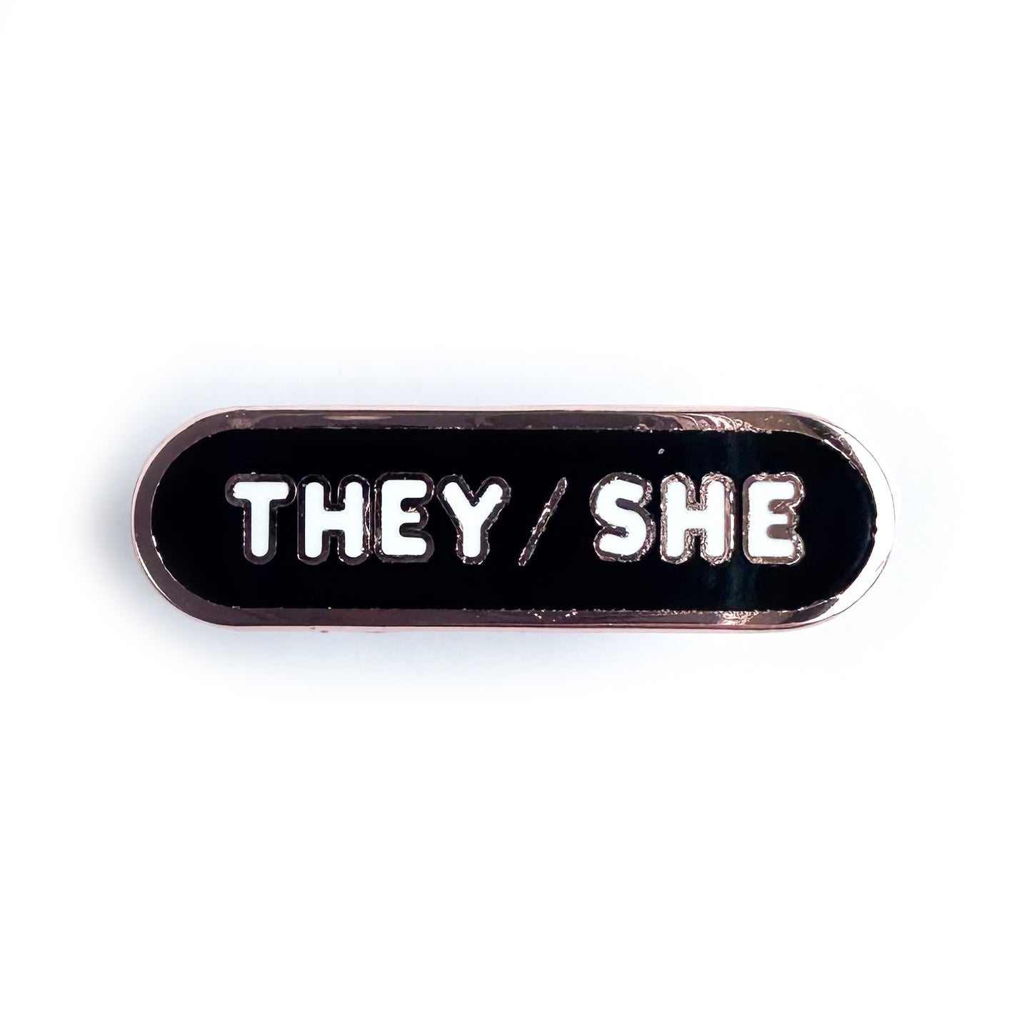 A black capsule shaped enamel pin with copper metal. The pin has white bubble letters that read "They/She" on it. 