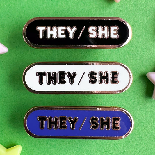 Three capsule shaped enamel pins with bubble letters that spell the pronouns "They/She", the top pin is black, the middle is white, and the bottom is indigo. The pins are on a green background.