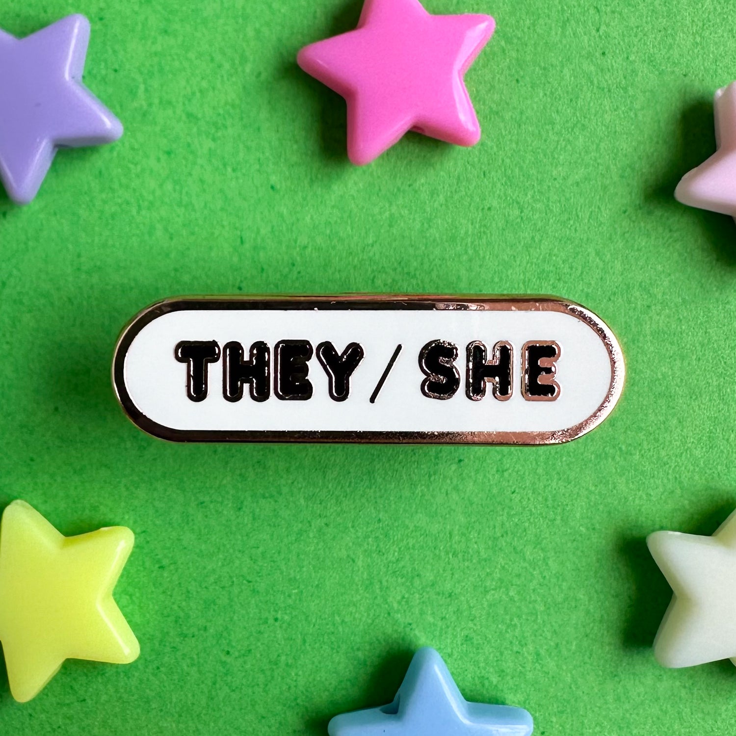 A white capsule shaped enamel pin with the pronouns "They/She' on it in black bubble letters. The pin is on a green paper background with plastic star beads around it. 