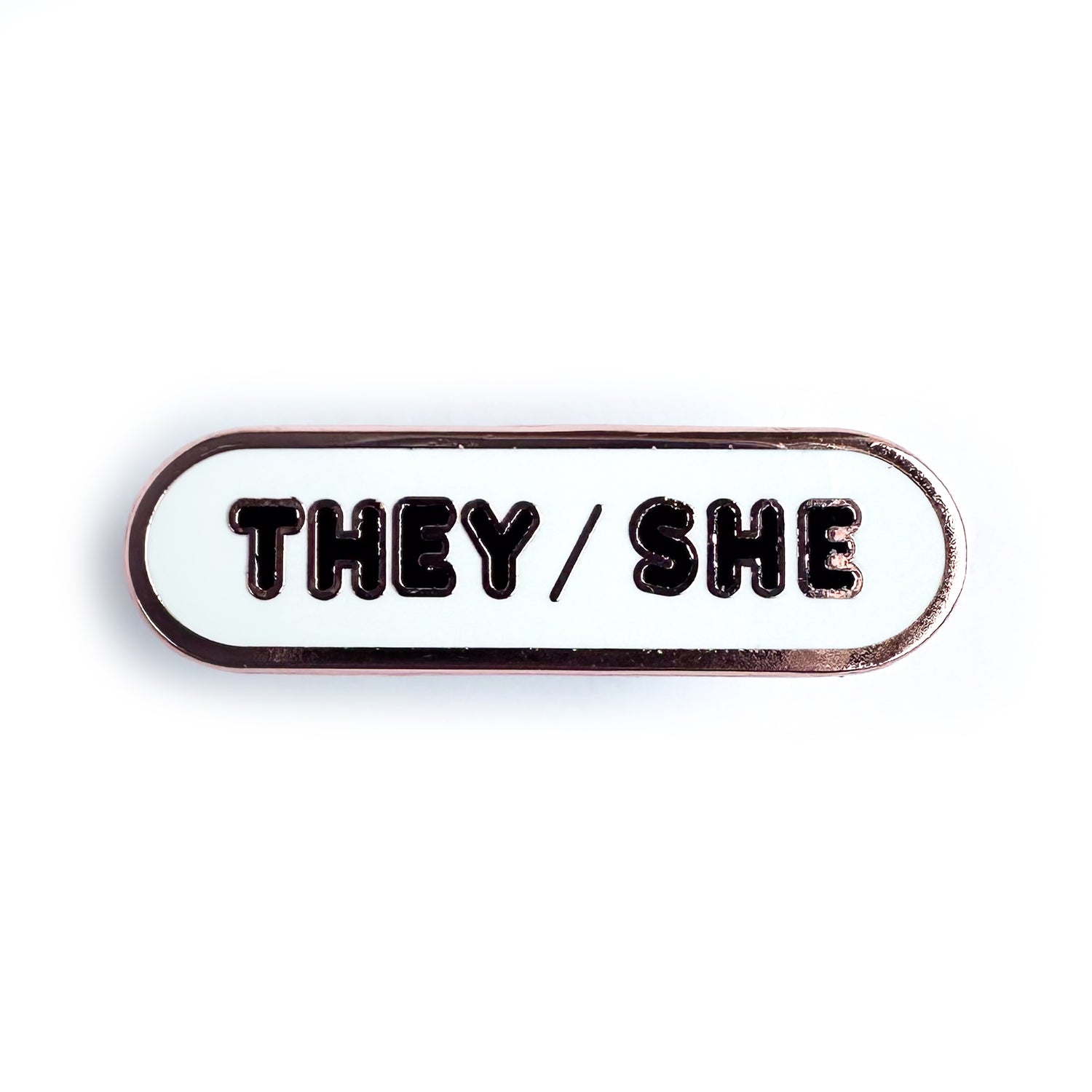 A white bandaid shaped enamel pin with black bubble letters that spell "They/She" on it. 