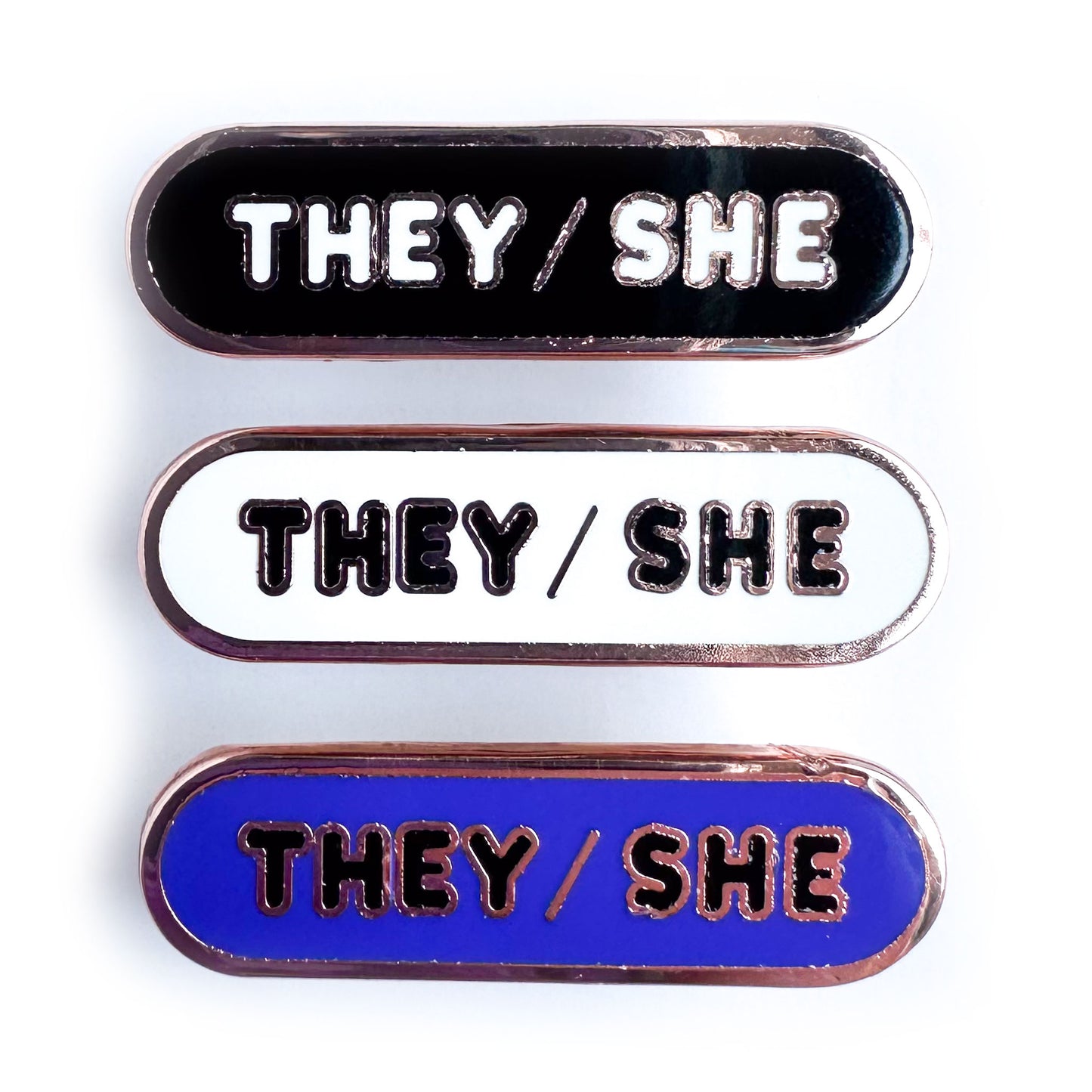 Three capsule shaped enamel pins with bubble letters that spell the pronouns "They/She", the top pin is black, the middle is white, and the bottom is indigo.