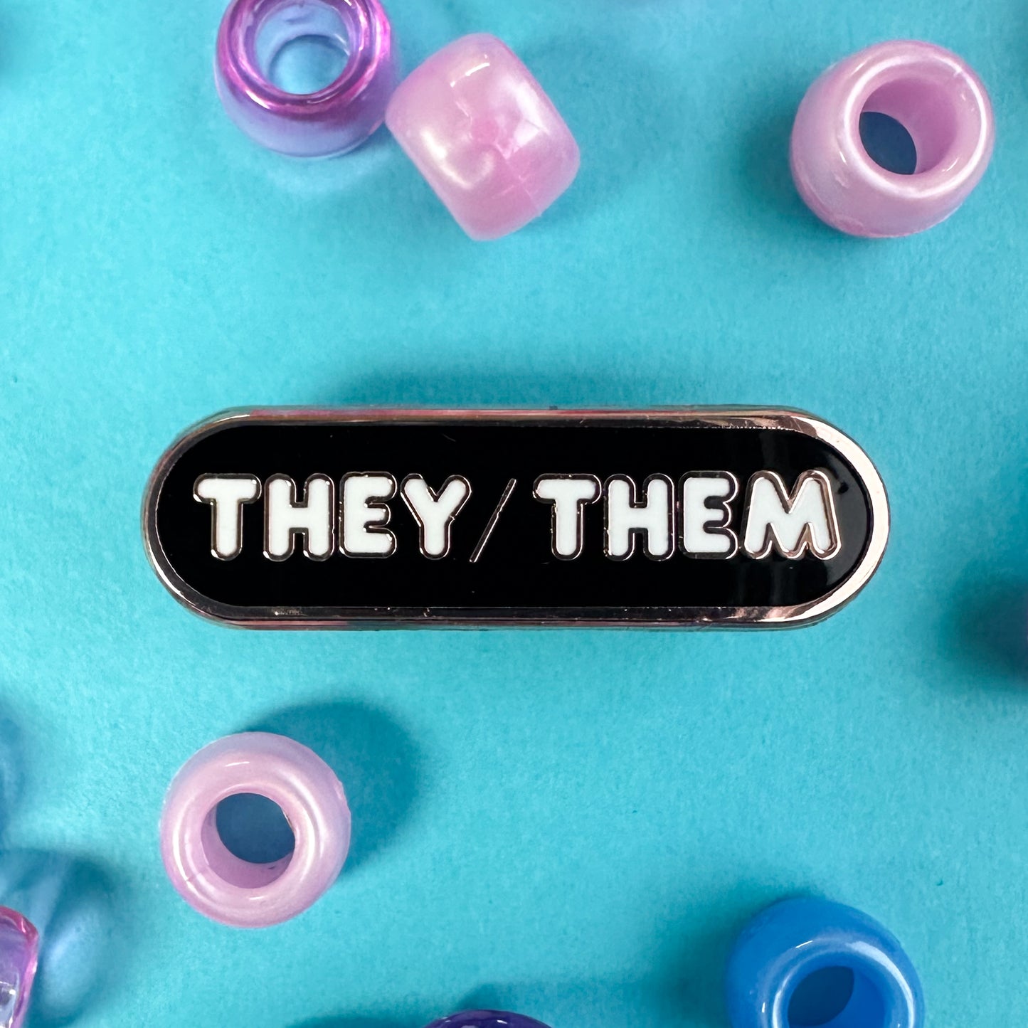 A black oval pin with the pronouns "They/Them" written on it in white bubble letters. The pin is on a blue background with pink and blue pony beads around them. 
