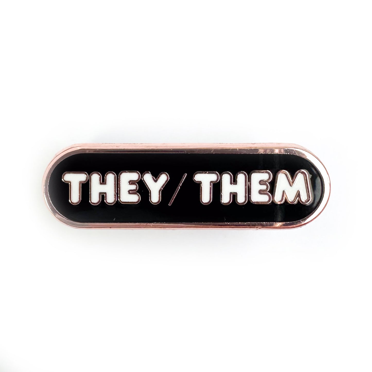 A black capsule shaped pin with white bubble letters that spell "They/Them" on it. 