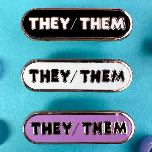 Three pins with the pronouns "They/Them" written on them, one pins is black, one is white, and one is lavender. The pins are on a blue background with blue pony beads around it. 