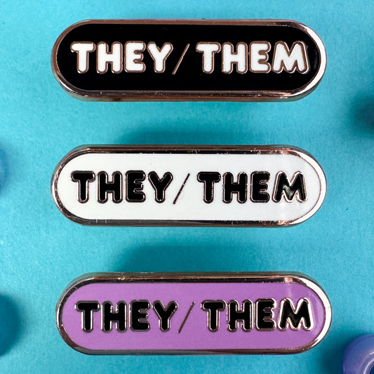 Three pins with the pronouns "They/Them" written on them, one pins is black, one is white, and one is lavender. The pins are on a blue background with blue pony beads around it.