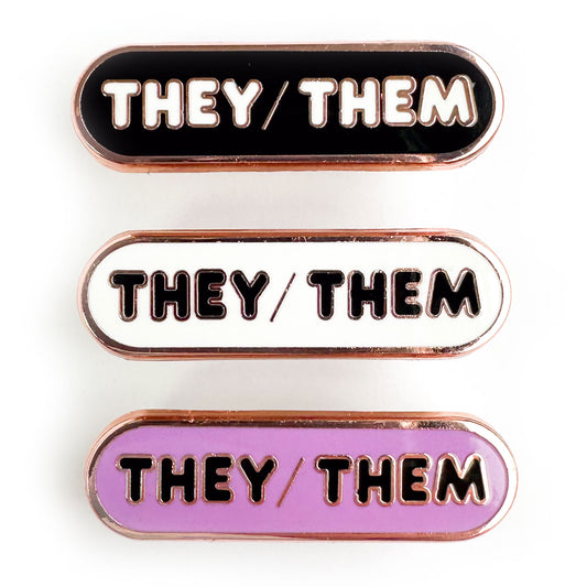Three capsule shaped pins that all have the words "They/Them" on them, one pin is black, one is white, and one is lavender. 