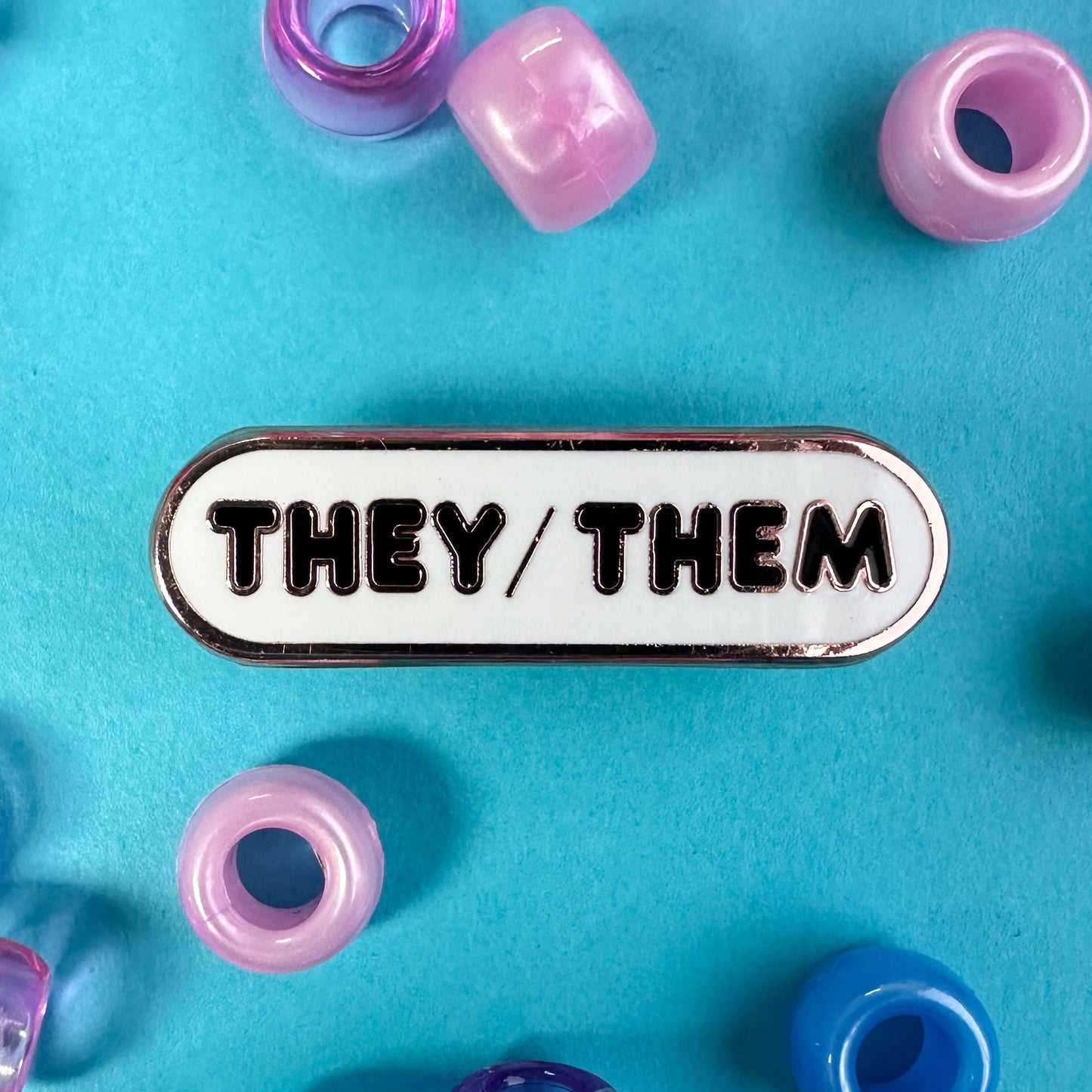 A bandaid shaped white pin with black bubble letters that spell "They/Them" on it. There are pink and blue pony beads scattered around it. 