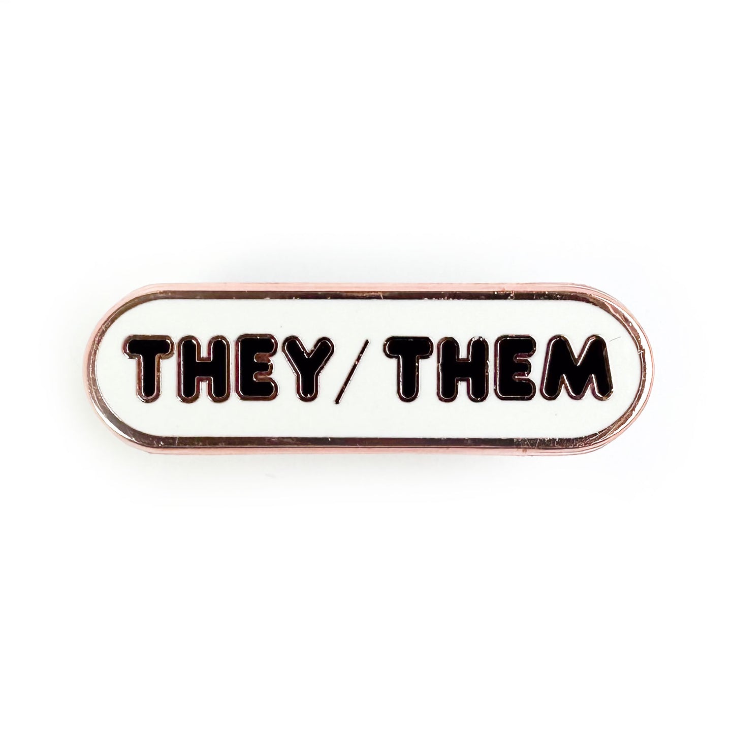 An oval shaped pin with a white background and black bubble letters on it, the letters spell "They/Them" on it. 