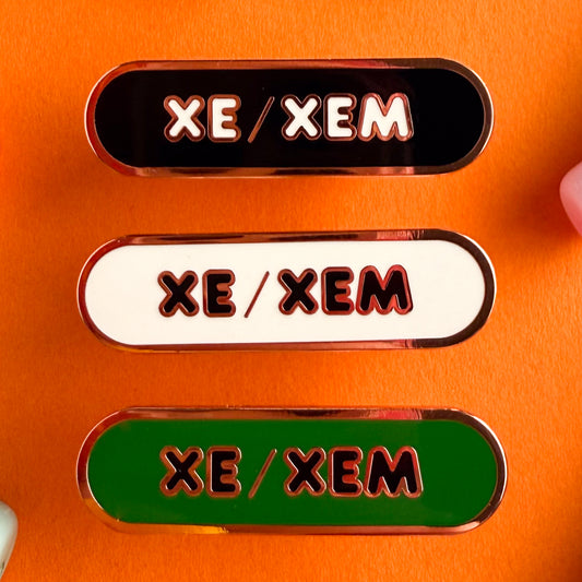 Three bandaid shaped pins with the text XE/XEM in bubble letters. Top one black with white text, the next is white with black text, and the last is green with black text. The pins are on an orange background.