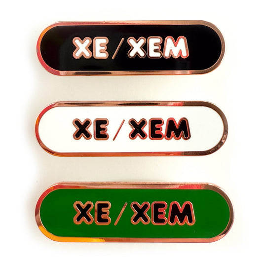 Three bandaid shaped pins with the text XE/XEM in bubble letters. Top one black with white text, the next is white with black text, and the last is green with black text.