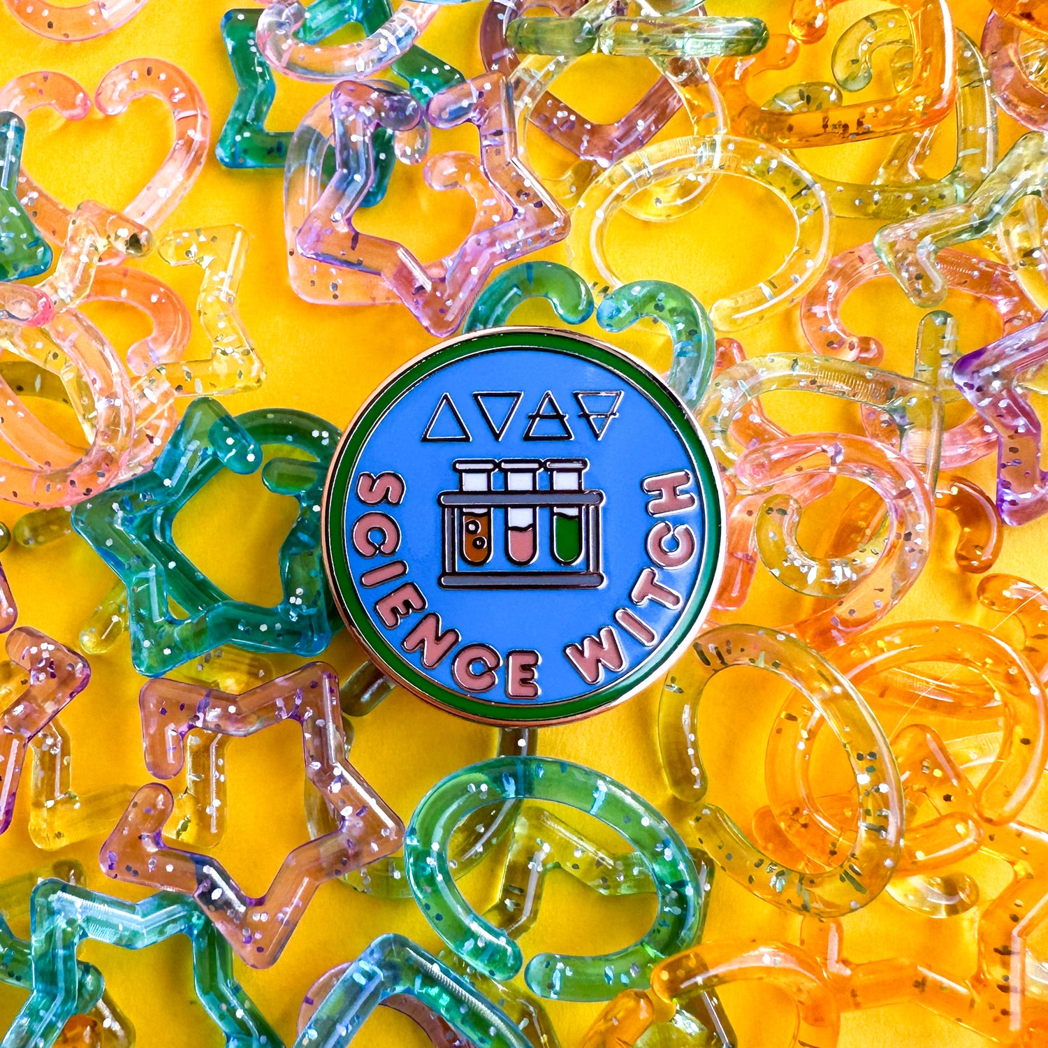 A circular pin that reads "Science Witch" with images of test tubes and the alchemical symbols for the four elements on it. The background of the pin is blue with a green border and the words are peach. The pin is on a yellow background with plastic glitter beads around it. 