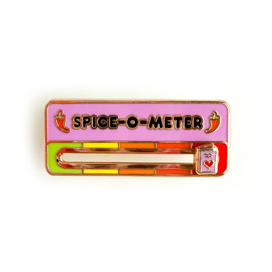 An enamel pin with the title SPICE-O-METER written in black on a pink background.There are two red hot peppers, one before and one after the title. Below the title is a scale with a slider in the middle. The scale is green, yellow, orange, red orange, and red. The slider is a small pink book with a red heart on the cover.