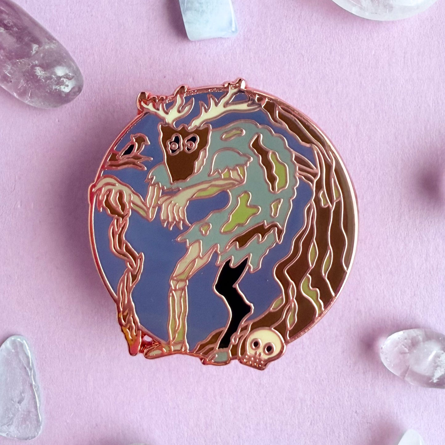 An enamel pin in the shape of a circle with a hunched zombie like creature with a goat leg and a mask with antlers standing near a tree and a skull. There is a bird in a brand next to the creature. The pin is on a purple background with small crystals strewn around it. 