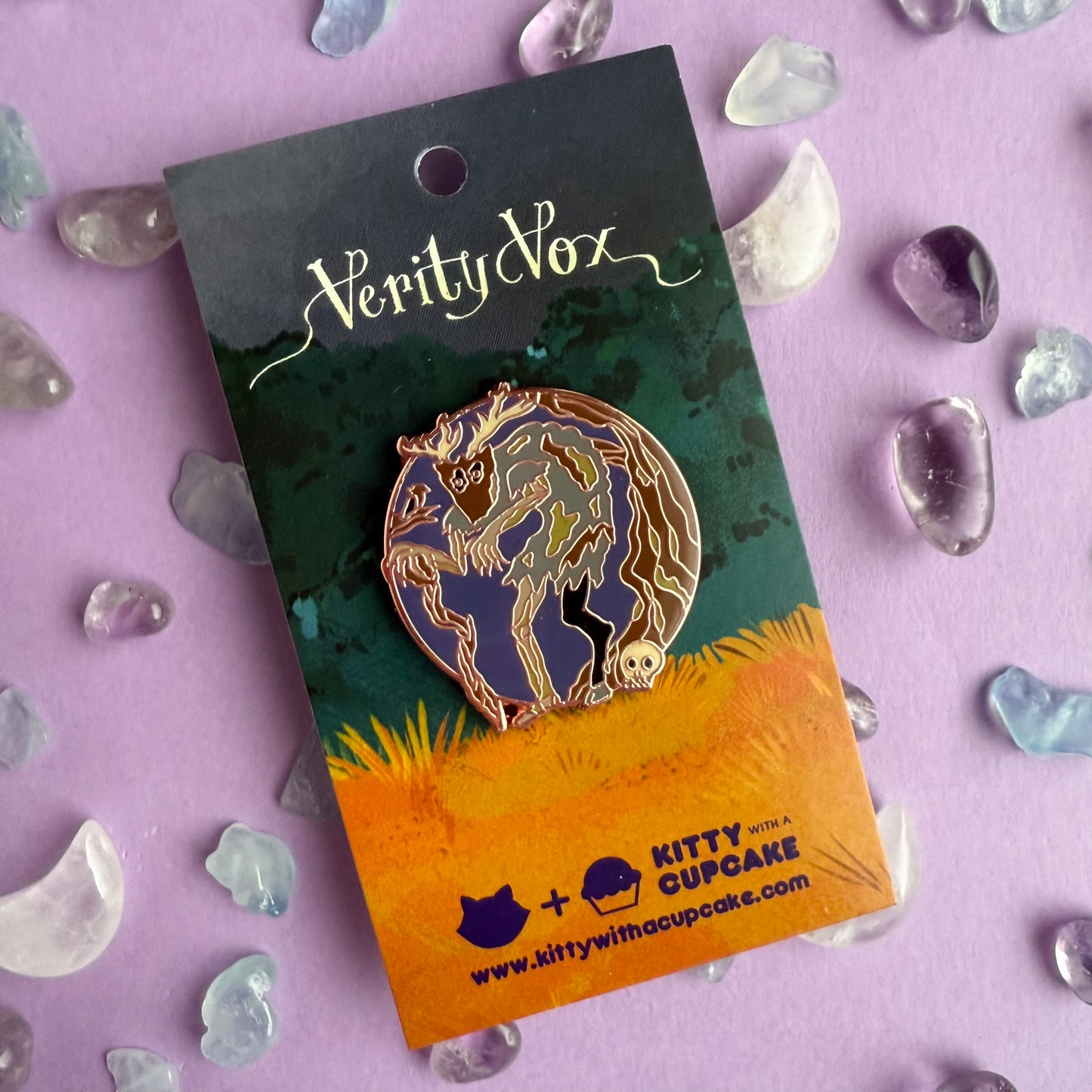 An enamel pin in the shape of a circle with a hunched zombie like creature with a goat leg and a mask with antlers standing near a tree and a skull. There is a bird in a brand next to the creature. The pin packaged on a card with the words Verity Vox in script font at the top and the Kitty With A Cupcake Logo on the bottom. There are crystals strewn about the card. 