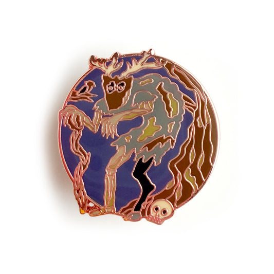 An enamel pin in the shape of a circle with a hunched zombie like creature with a goat leg and a mask with antlers standing near a tree and a skull. There is a bird in a brand next to the creature. 