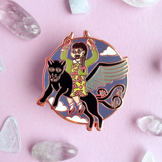 An enamel pin in the shape of a circle with an illustration of a woman holding knitting needles up like daggers wearing a cardigan and a floral dress. The woman is riding a winged jaguar with barbed tails flying through the sky. The pin is on a lavender background with small crystals around it.