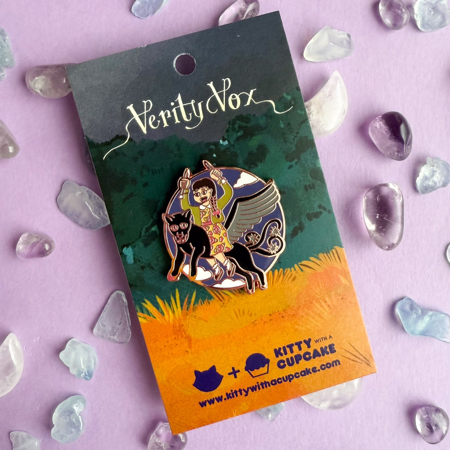 An enamel pin in the shape of a circle with an illustration of a woman holding knitting needles up like daggers wearing a cardigan and a floral dress. The woman is riding a winged jaguar with barbed tails flying through the sky. The pin is packaged on a card with scripty font reading "Verity Vox" at the top and the Kitty With A Cupcake logo at the bottom. 