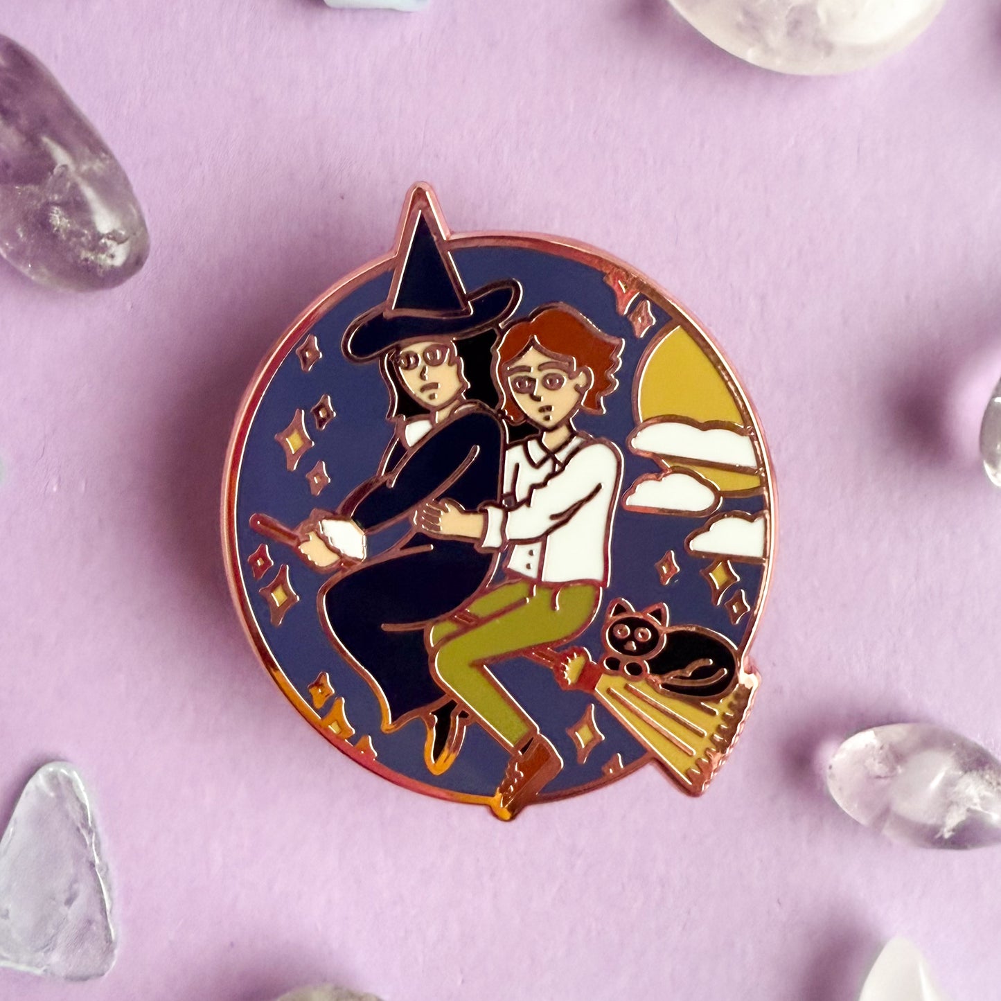 An enamel pin featuring Verity and Tacita flying away together on Verity's broom. Verity's cat-who-is-not-a-cat familiar, Jack is riding with them! There are stars, clouds, and the moon surrounding them in a circle. The pin is on a purple paper background with small crystals strewn about.