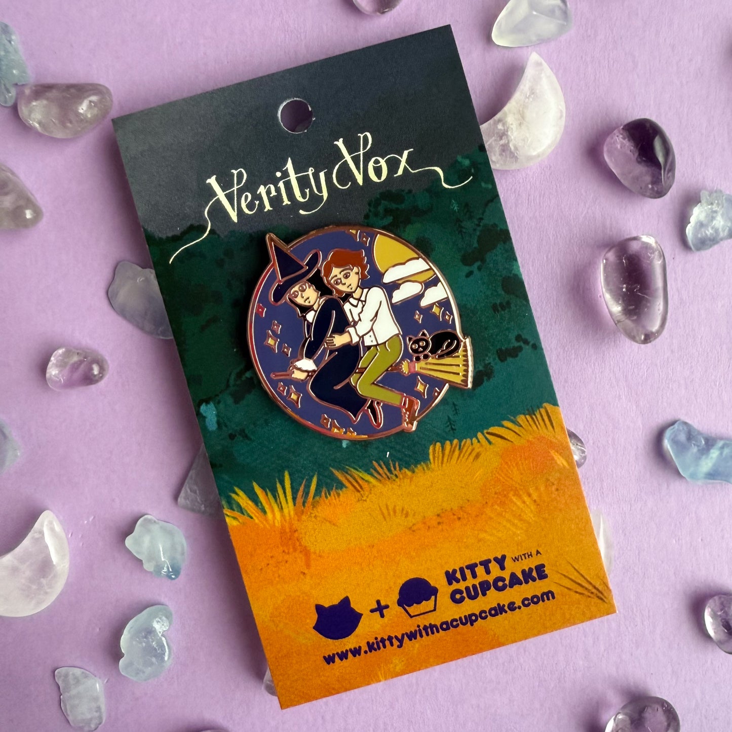 An enamel pin featuring Verity and Tacita flying away together on Verity's broom. Verity's cat-who-is-not-a-cat familiar, Jack is riding with them! There are stars, clouds, and the moon surrounding them in a circle. It is packaged on a card with the words Verity Vox in a script font and the Kitty With A Cupcake Logo. The pin is on a purple paper background with small crystals strewn about. 