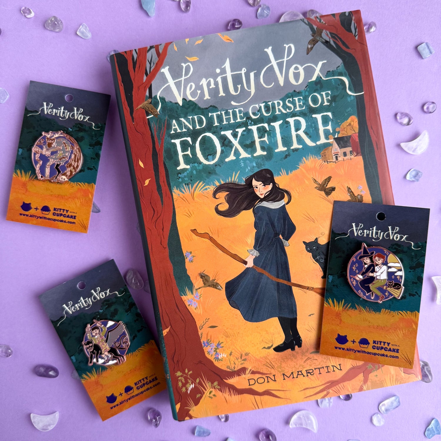 A hardcover copy of Verity Vox and the Curse Foxfire with three pins with packaging matching the gold and teal colors of the book. 