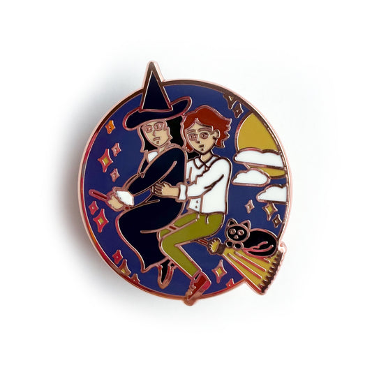 An enamel pin featuring Verity and Tacita flying away together on Verity's broom. Verity's cat-who-is-not-a-cat familiar, Jack is riding with them! There are stars, clouds, and the moon surrounding them in a circle.