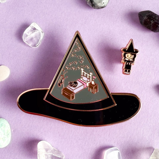 An enamel pin set, one is a large pin shaped like a black witch hat with illustrations of a bedroom set and a cat in it. The other pin is a mini pin of a witch wearing a hat that matches the other hat. The pins are surrounded by small crystals and are on a light purple background.