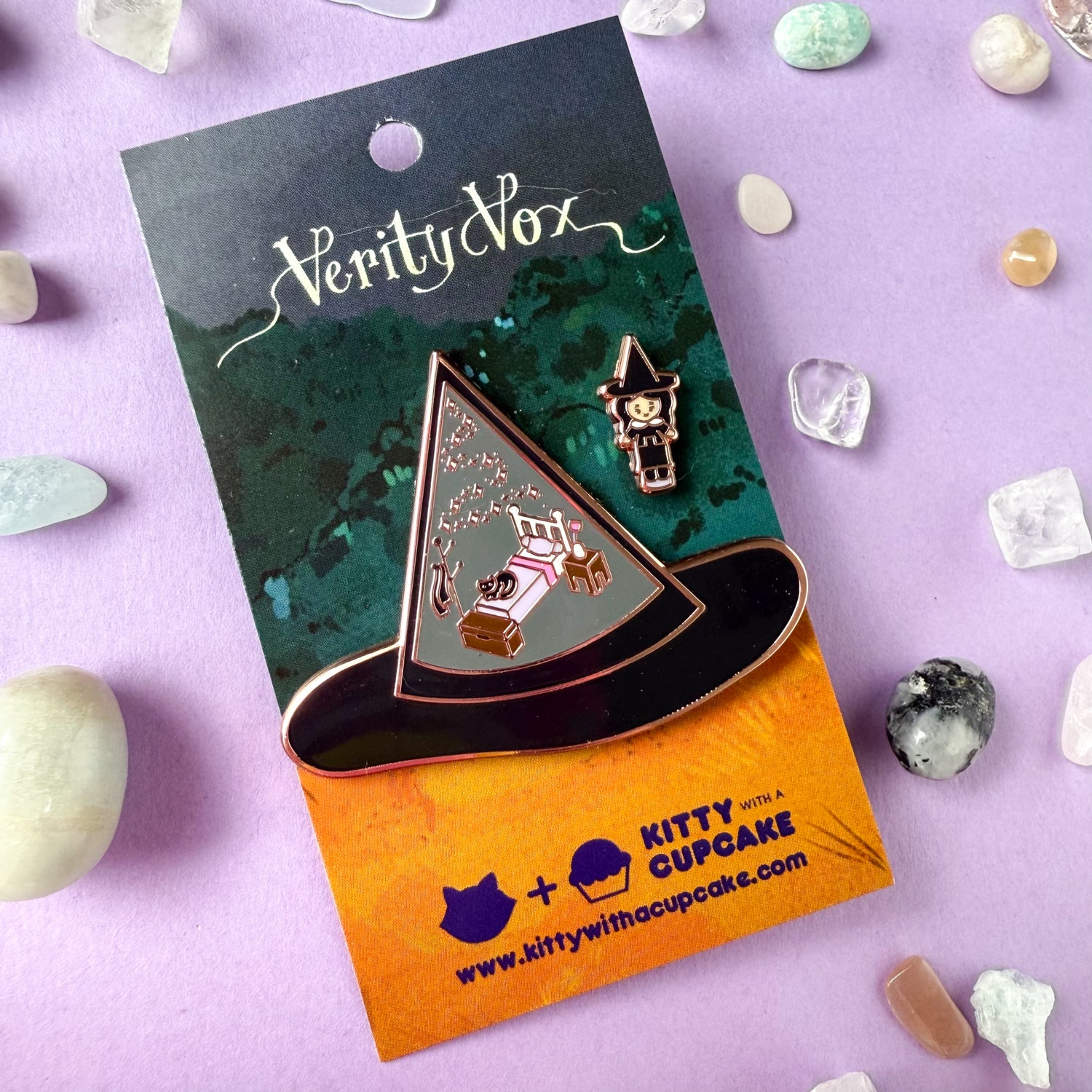 An enamel pin set, one is a large pin shaped like a witch hat with illustrations of a bedroom set and a cat in it. The other pin is a mini pin of a witch wearing a hat that matches the other hat.  The pins are packaged on a Verity Vox branded card and are surrounded by small crystals. 