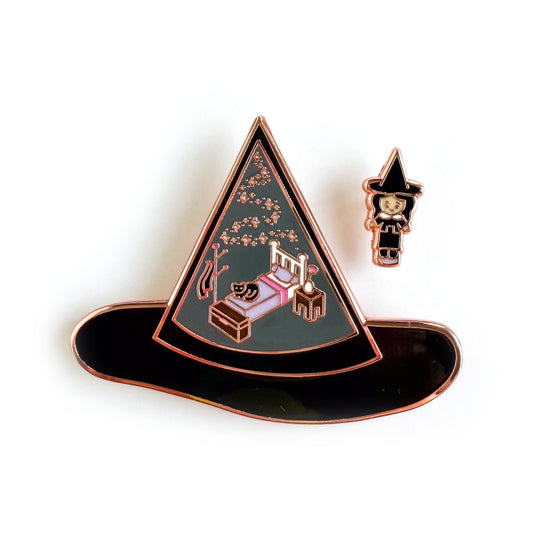 An enamel pin set, one is a large pin shaped like a witch hat with illustrations of a bedroom set and a cat in it. The other pin is a mini pin of a witch wearing a hat that matches the other hat.