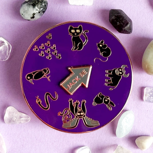 A round purple enamel pin with illustrations of black animals in a circle. There is a black cat, mouse, bull, rabbit, chimera, snake, raven, and swarm of flies. There is a spinning arrow in the middle of the circle that reads "Jack Be". There are small crystals around the pin.