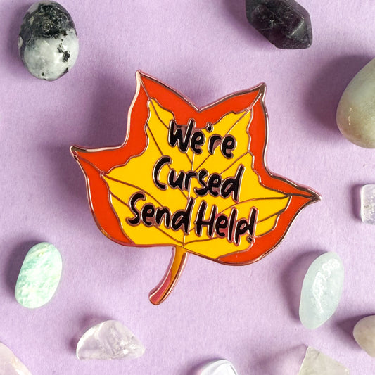 A pin shaped like a tulip poplar leaf in fall colors with the words "We're cursed send help!" written on it. The pin is surrounded by small crystals and is on a light purple background.