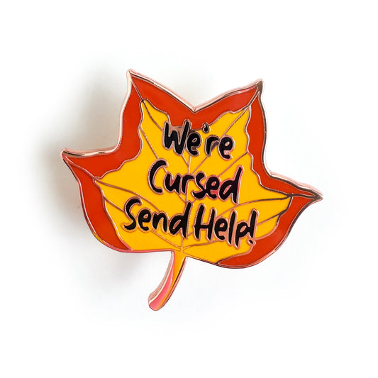 A pin shaped like a tulip poplar leaf in fall colors with the words "We're cursed send help!" written on it. 