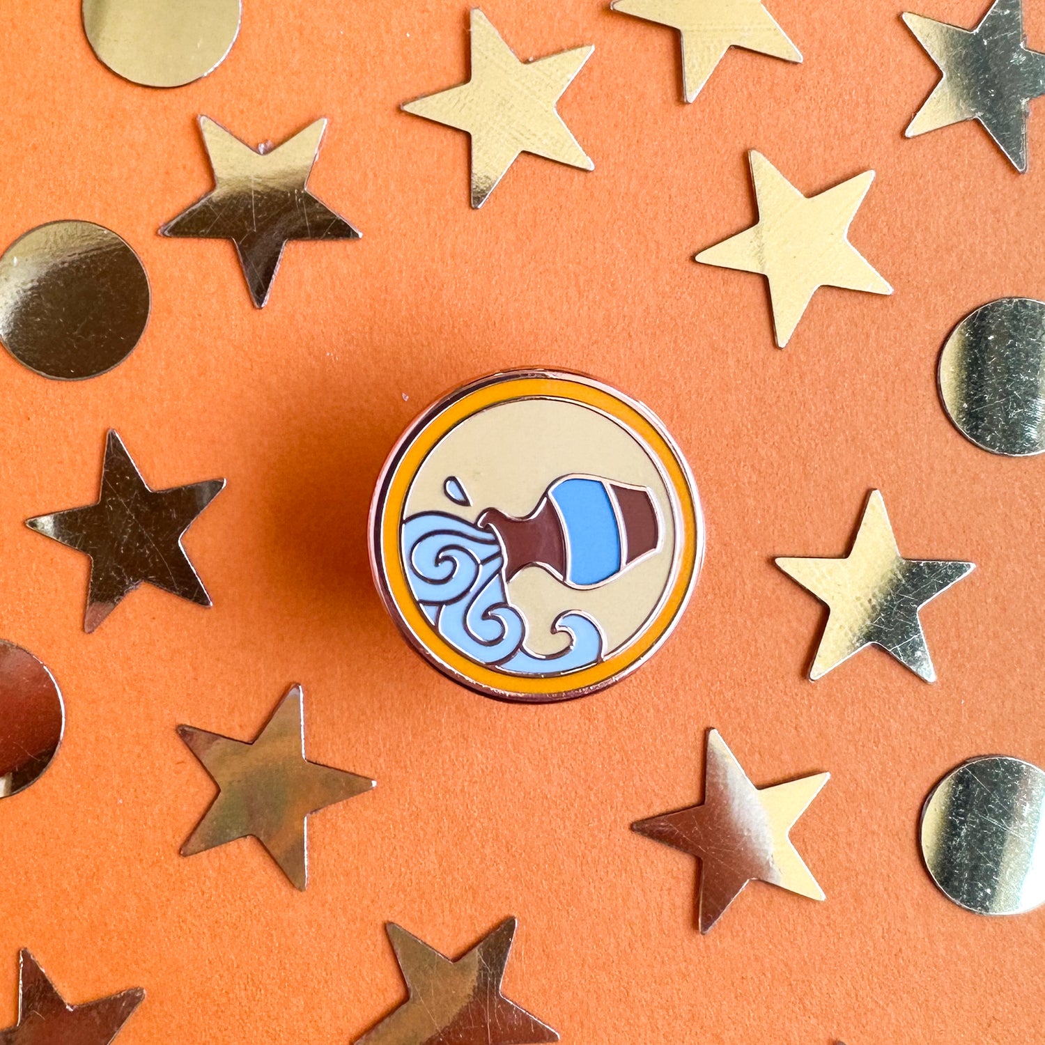 A circular pin containing a cute version of the Aquarius symbol, a water vessel pouring water, on an orange background with glitter stars and circles surrounding it. 