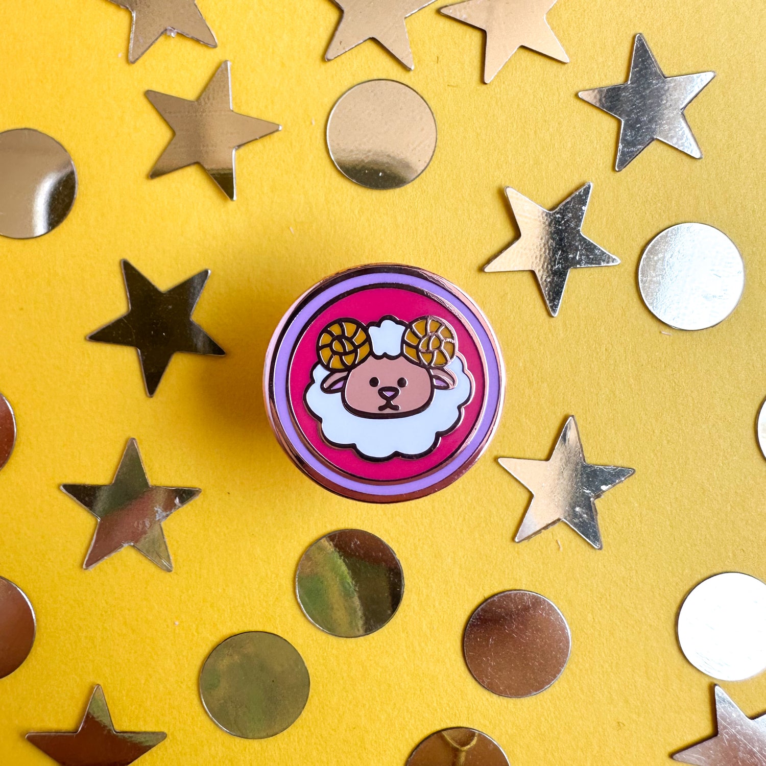 A circular enamel pin with a cute sheep on it sitting on a yellow background with gold glitter stars and circles around it. 
