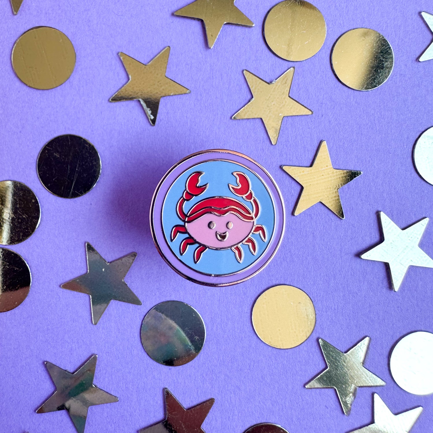 An enamel pin in the shape of a circle with a cute little crab illustration inside of it to represent Cancer in the zodiac. The pin is on a purple background with gold stars and circles around it. 