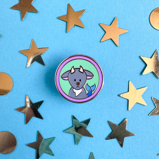 An enamel pin in the shape of a circle with a cute sea goat illustration to represent the Capricorn zodiac sign. The pin is on a blue paper background with gold stars and circles around it. 