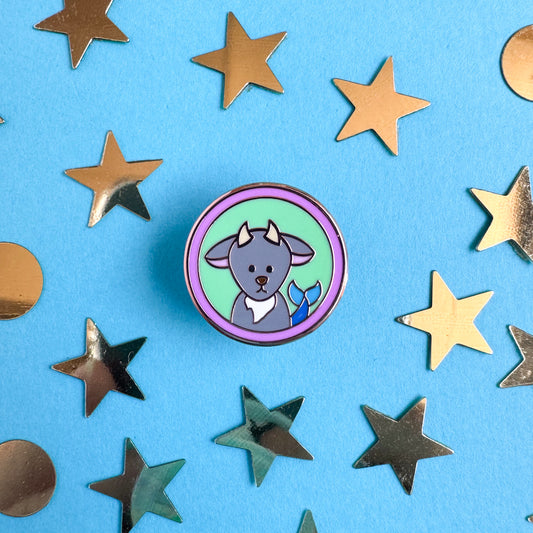 An enamel pin in the shape of a circle with a cute sea goat illustration to represent the Capricorn zodiac sign. The pin is on a blue paper background with gold stars and circles around it.