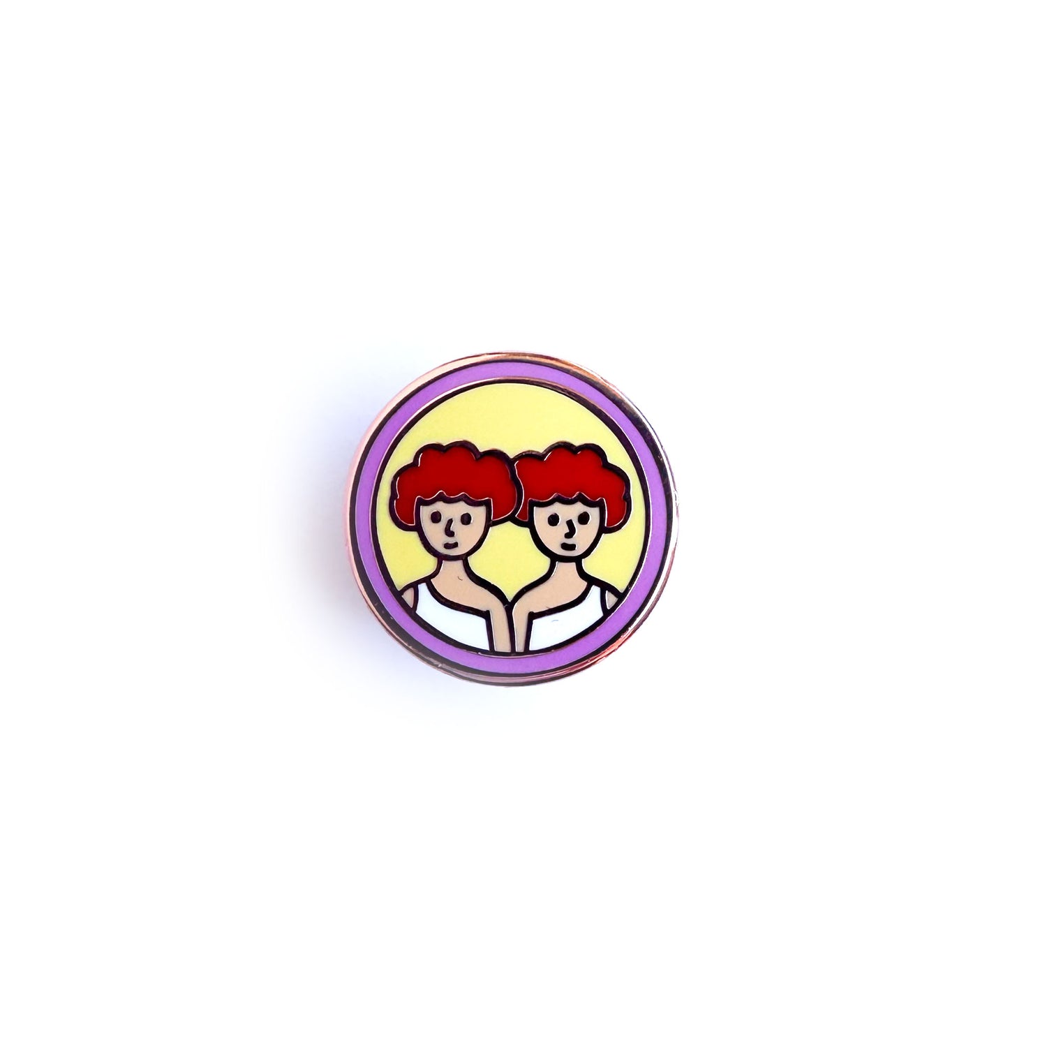 An enamel pin in the shape of a circle with art of two twins with read hair on it to represent the Gemini Zodiac sign. 