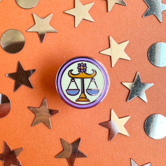 A circle enamel pin with scales on it to represent the Libra zodiac sign. The pin is on an orange background that is covered with gold stars and circle confetti.