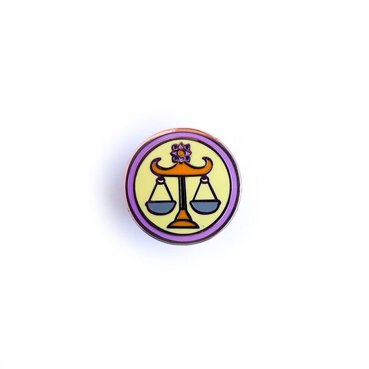 A circular enamel pin with the symbol for Libra, scales of balance.