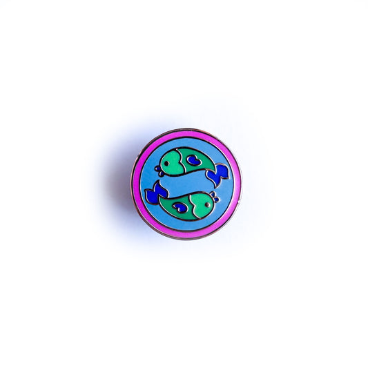A pin of a blue circle with a border of a hot pink circle with two green fish swimming in opposite directions on the blue circle to represent the zodiac symbol of Pisces.