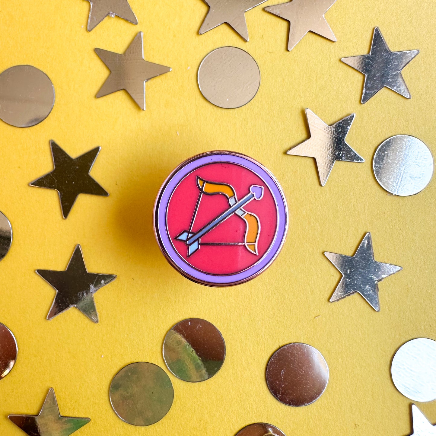 A circle enamel pin with a cute pink bow and arrow in it to represent Sagittarius. The pin is on a yellow background with gold confetti around it. 